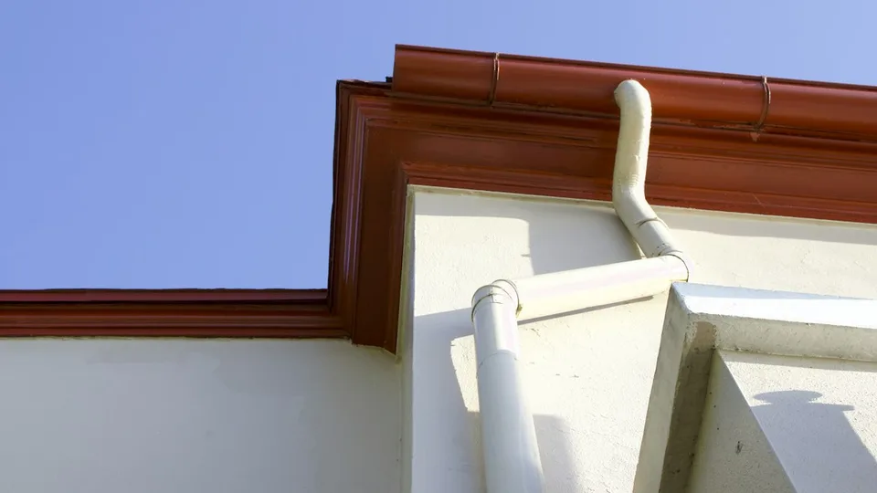Sarkana jumta tekne un balta notekcaurule pie gaišas fasādes