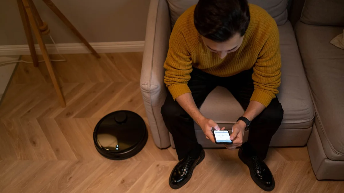 Cilvēks viesistabā lieto telefonu līdzās robotam putekļsūcējam